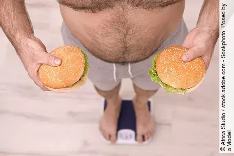 Übergewichtiger auf der Waage, in beiden Händen ein Burger