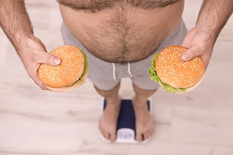 Übergewichtiger auf der Waage, in beiden Händen ein Burger