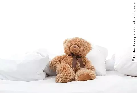 Teddybär sitzt auf einem Bett mit weißen Laken.