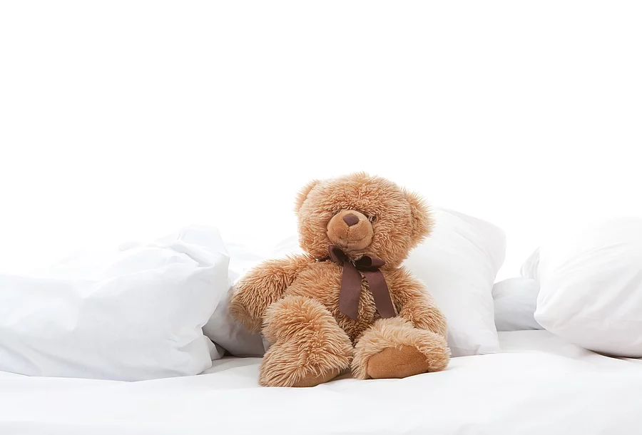Teddybär sitzt auf einem Bett mit weißen Laken.