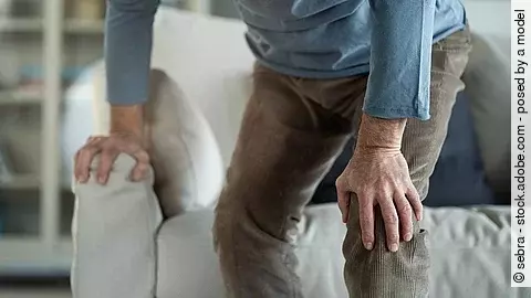 Älterer Mann steht hält sich beim Aufstehen vom Sofa das schmerzende Knie