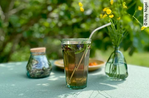 Ein Glas mit Tee, Kräutern und Strohhalm; daneben ein Blumenstrauß und eine Dose; alles auf einem Tisch in einem Garten