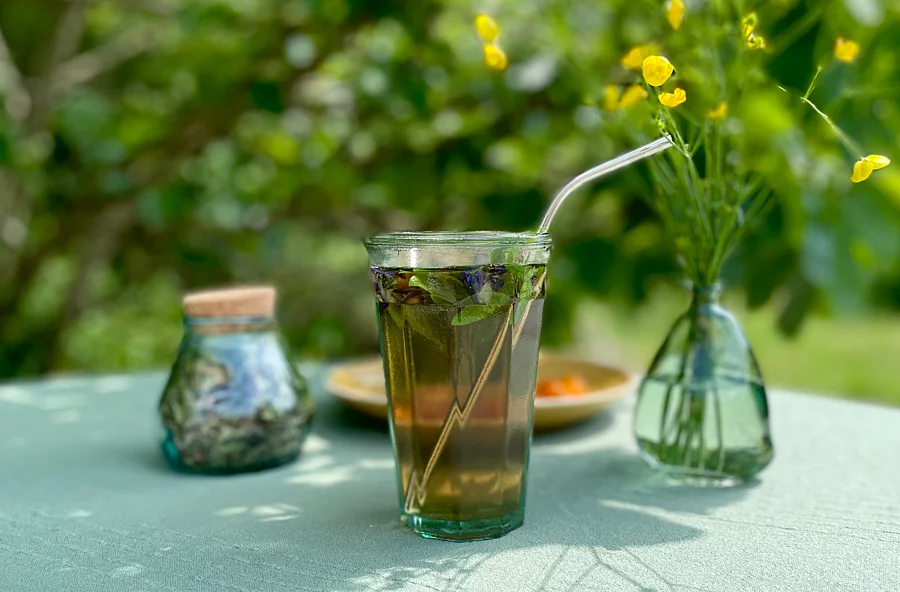 Ein Glas mit Tee, Kräutern und Strohhalm; daneben ein Blumenstrauß und eine Dose; alles auf einem Tisch in einem Garten