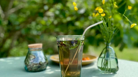 Ein Glas mit Tee, Kräutern und Strohhalm; daneben ein Blumenstrauß und eine Dose; alles auf einem Tisch in einem Garten
