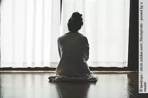 Frau sitzt auf dem Boden vor Fenster mit geschlossenen Gardinen