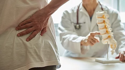Eine Frau hält sich mit ihrer rechten Hand den unteren Rücken, im Hintergrund sitzt ein Arzt an einem Tisch.