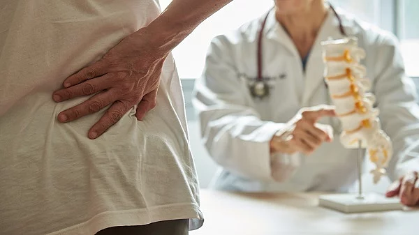 Eine Frau hält sich mit ihrer rechten Hand den unteren Rücken, im Hintergrund sitzt ein Arzt an einem Tisch.