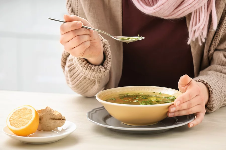 Sick young woman eating broth to cure cold at table in kitchen Frau isst eine Hühnersuppe.