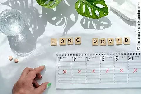 Holzplättchen, die das Wort Long-COVID bilden, darunter ein Kalender und eine Hand.