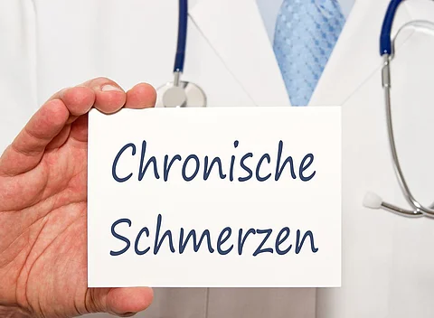 Ein Arzt im Kittel und mit Stethoskop um den Hals hält ein kleines Schild mit der Aufschrift "Chronische Schmerzen" in der rechten Hand