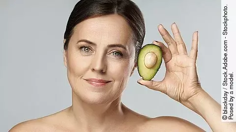 Frontalansicht von Frau die eine Avocado neben ihr Gesicht hält.