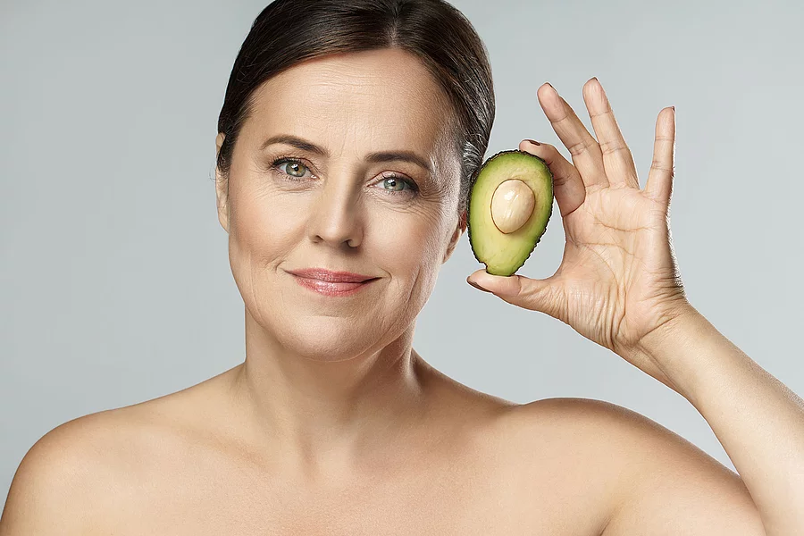 Frontalansicht von Frau die eine Avocado neben ihr Gesicht hält.