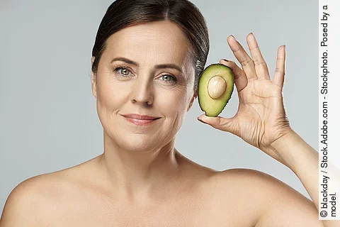 Frontalansicht von Frau die eine Avocado neben ihr Gesicht hält.