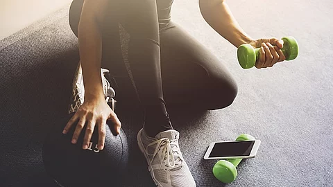 Frau benutzt Hanteln beim Sport. Vor ihr liegt ein Smartphone. 