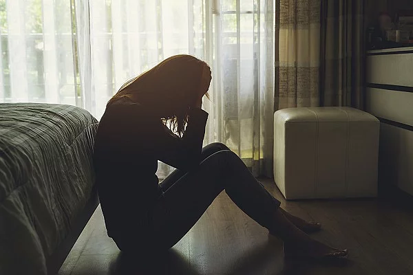 Eine depressive Frau sitzt auf dem Boden in einem dunklen Schlafzimmer und fasst sich mit ihren Händen an den Kopf.