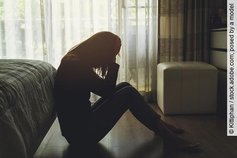 Eine depressive Frau sitzt auf dem Boden in einem dunklen Schlafzimmer und fasst sich mit ihren Händen an den Kopf.