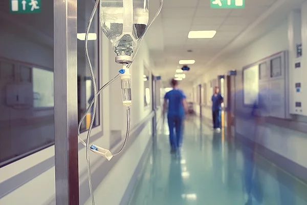 Tropf steht in einem Krankenhaus-Korridor. Im Hintergrund sind Pflegekräfte zu sehen.