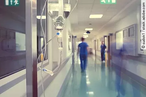 Tropf steht in einem Krankenhaus-Korridor. Im Hintergrund sind Pflegekräfte zu sehen.