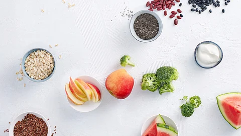 Tableau mit ballaststoffreichen Nahrungsmitteln: Leinsamen, Apfel, Broccoli, Melone, Haferflocken, Chia, rote und schwarze Bohnen