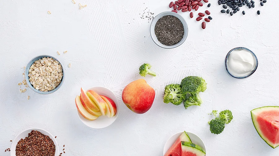 Tableau mit ballaststoffreichen Nahrungsmitteln: Leinsamen, Apfel, Broccoli, Melone, Haferflocken, Chia, rote und schwarze Bohnen