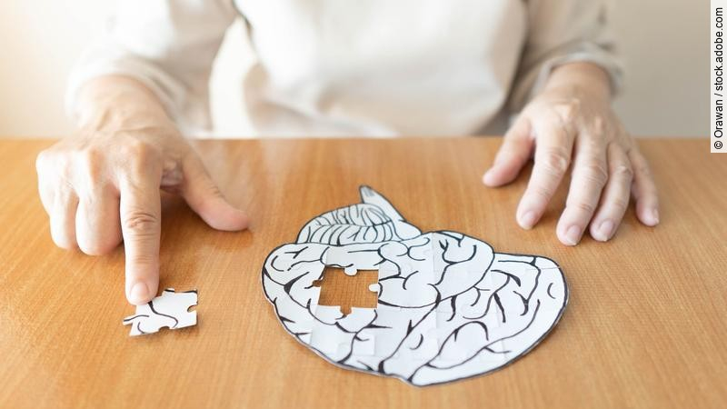 Hochbetagte Frau sitzt vor einem Puzzle in Form eines Gehirns, bei dem ein Teil fehlt.