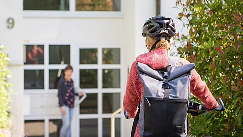 Frau mit Fahrrad vor einem Wohnhaus, an der Tür wartet eine andere Frau auf sie.