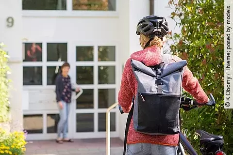 Frau mit Fahrrad vor einem Wohnhaus, an der Tür wartet eine andere Frau auf sie.