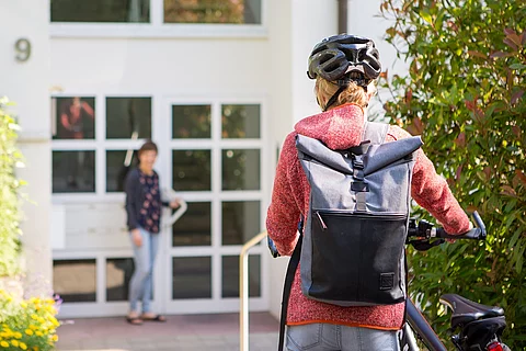 Frau mit Fahrrad vor einem Wohnhaus, an der Tür wartet eine andere Frau auf sie.