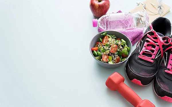 Rechts im Bild befinden sich Fitness-Equipment, ein Salat, eine Wasserflasche, ein Apfel und Kopfhörer.