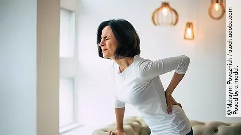 Frau mittleren Alters steht mit schmerzverzerrtem Gesicht im Wohnzimmer mit einer Hand am Rücken und der anderen abgestützt auf der Couch.