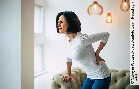 Frau mittleren Alters steht mit schmerzverzerrtem Gesicht im Wohnzimmer mit einer Hand am Rücken und der anderen abgestützt auf der Couch.