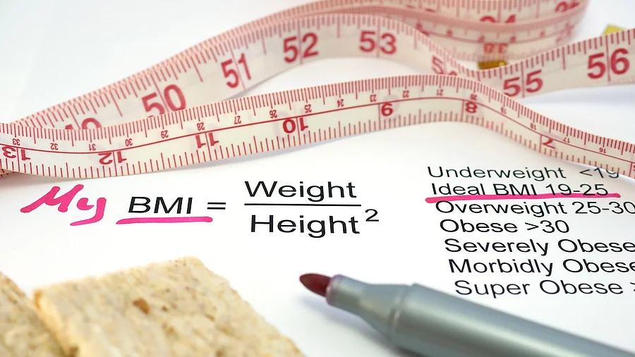 Auf einem Schreibtisch liegen ein Maßband, ein Zettel mit der Formel für den BMI und ein lila Filzstift. 