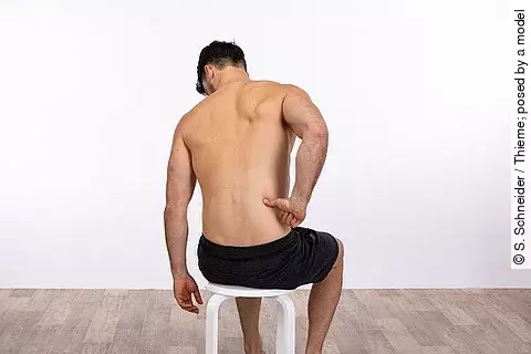 Mann mit nacktem Oberkörper, der auf einem weißen Hocker sitzt und sich mit der rechten Hand an den unteren Rücken fasst.