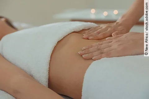 Frau liegt auf einer Liege mit Handtüchern bedeckt. Eine andere Frau legt die Hände auf den Bauch der Liegenden.