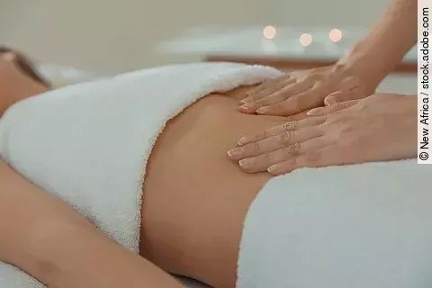 Frau liegt auf einer Liege mit Handtüchern bedeckt. Eine andere Frau legt die Hände auf den Bauch der Liegenden.