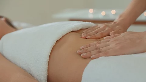 Frau liegt auf einer Liege mit Handtüchern bedeckt. Eine andere Frau legt die Hände auf den Bauch der Liegenden.