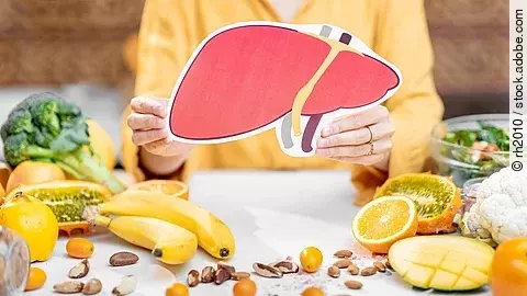 Frau hält Zeichnung einer Leber in der Hand. Auf dem Tisch liegen mediterrane Obst- und Gemüsesorten. 