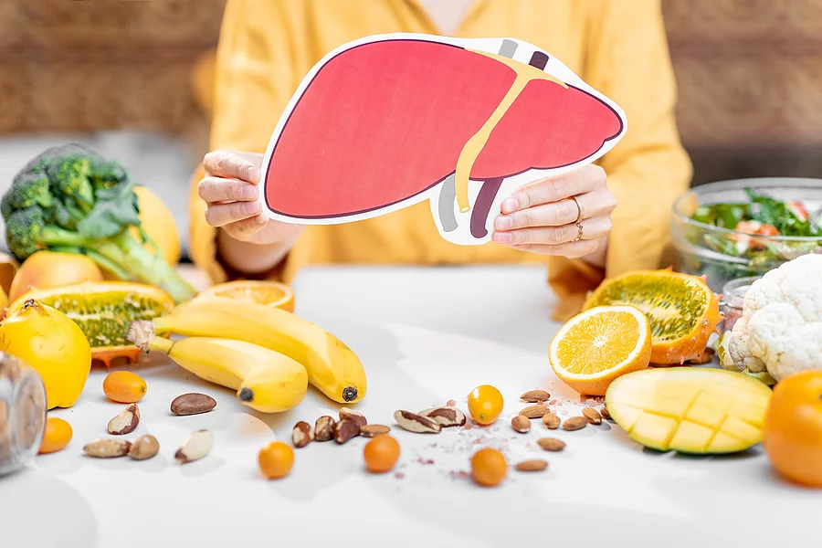 Frau hält Zeichnung einer Leber in der Hand. Auf dem Tisch liegen mediterrane Obst- und Gemüsesorten. 