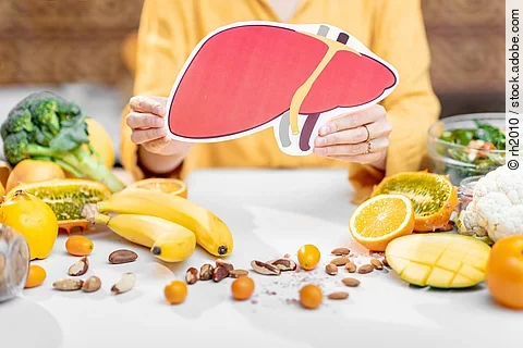 Frau hält Zeichnung einer Leber in der Hand. Auf dem Tisch liegen mediterrane Obst- und Gemüsesorten. 