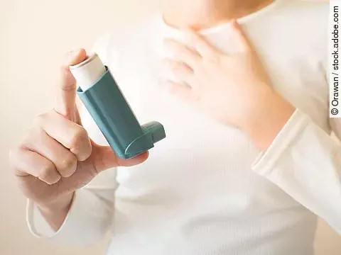 Ein Kind mit weißem Shirt hält in der einen Hand ein blaues Asthmaspray und fasst sich mit der anderen Hand an die Brust. 