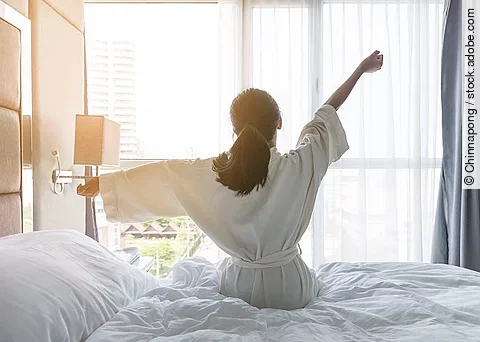 Frau sitzt auf dem Bett und streckt die Arme zur Seite. 