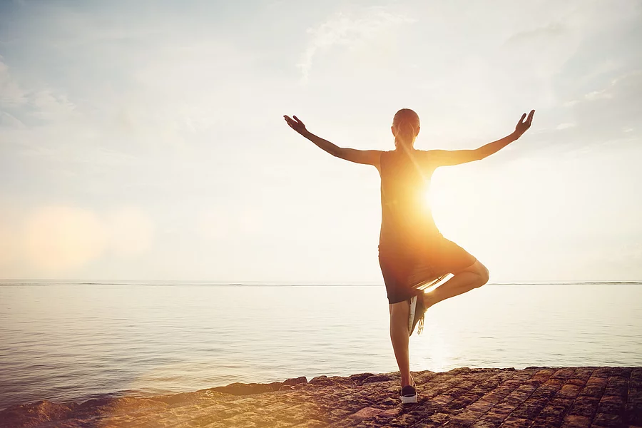 Young woman standing in yoga pose on the beach and meeting sunrise. Intentional sun glare, lens flare effect Frau in Baum-Asana an einem Strand