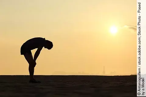 Silhouette eines gebeugt stehenden, erschöpften Sportlers vor Sonnenuntergang