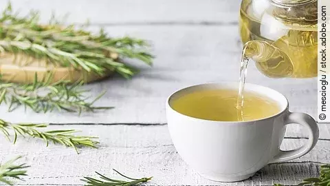 White cup of healthy rosemary tea pouring from teapot with fresh rosemary bunch on white wooden rustic background, winter herbal hot drink concept, salvia rosmarinus Teekanne und Teetasse, dekoriert mit frischen Teezweigen