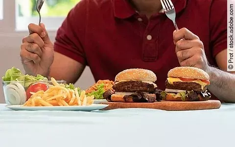 Mann mit Messer und Gabel am Tisch mit Burger, Pommes und Salat
