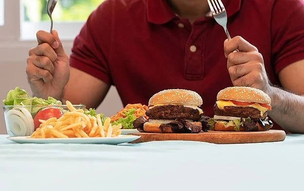 Young Man holding knife and fork are ready to eating a hamburger, French fries, and Spaghetti for Lunch. Concept of binge eating disorder (BED) and Relaxing with Eating junk food and unhealthy foods. Mann mit Messer und Gabel am Tisch mit Burger, Pommes und Salat