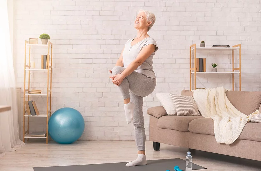 Seniorin macht Übungen auf einem Bein im Wohnzimmer