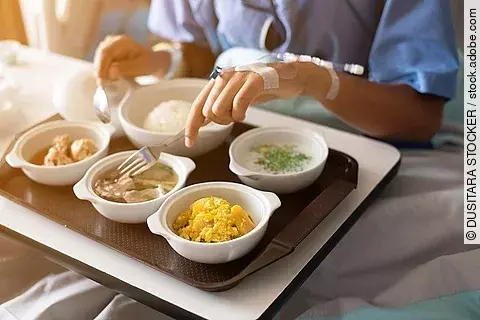 Patient liegt im Krankenbett mit einem Tablett voller Essen vor sich. 