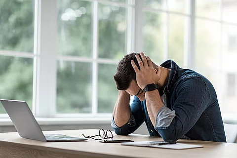Ein Mann sitzt an einem Schreibtisch und stützt den Kopf erschöpft auf seine Hände.