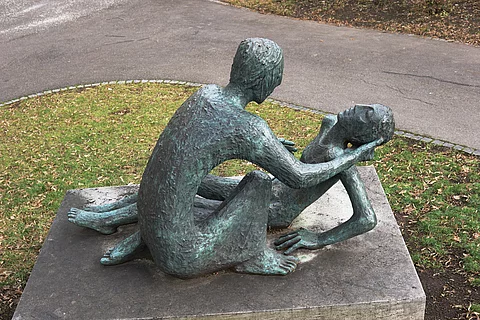 2 Skulpturen in einem Park: eine männliche Skultur beugt sich über eine auf dem Boden sitzende weibliche Skulptur